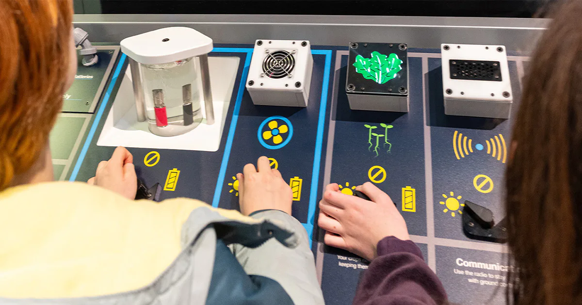 Two children interact with an educational exhibit featuring buttons and dials. The panel displays icons of plants, fans, and light bulbs, suggesting themes of energy or science.