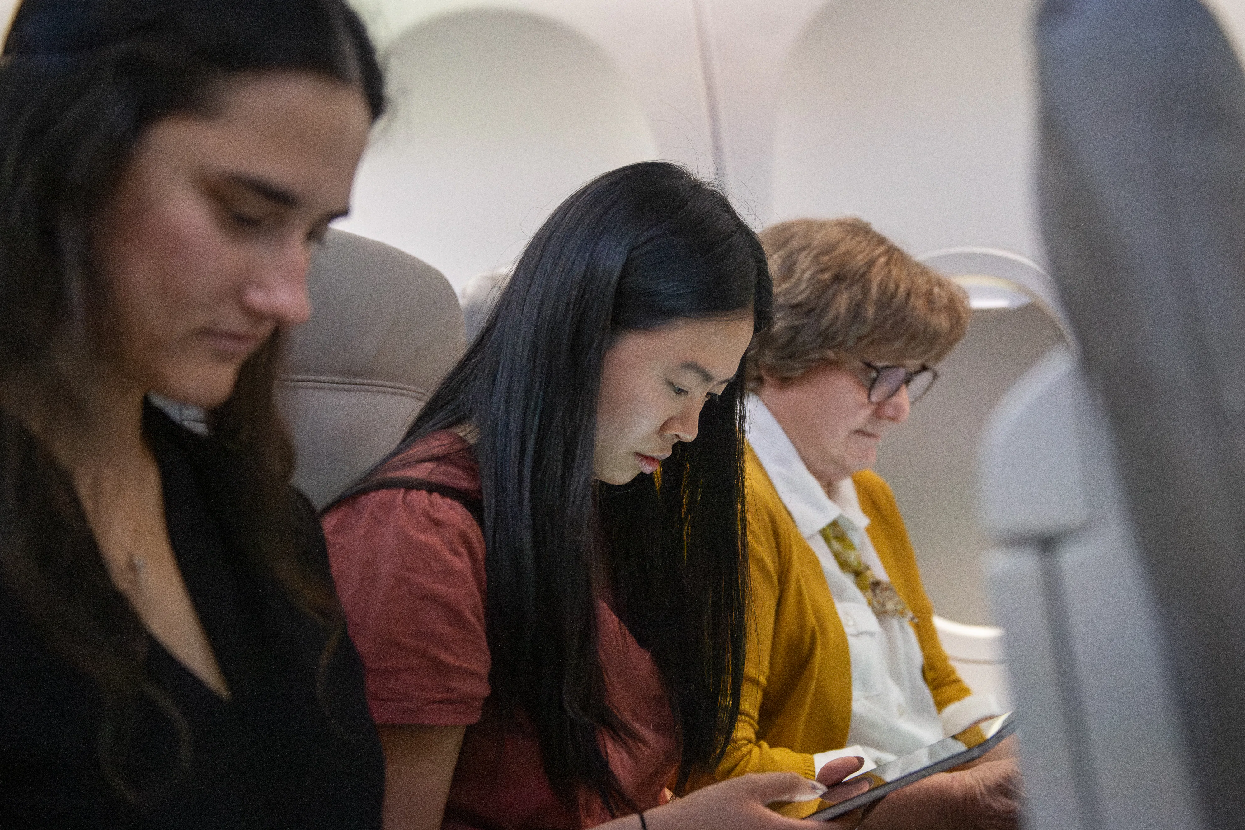 Three women seated in an airplane, focused on their devices. The mood is calm, and the airplane interior creates a quiet, introspective atmosphere.