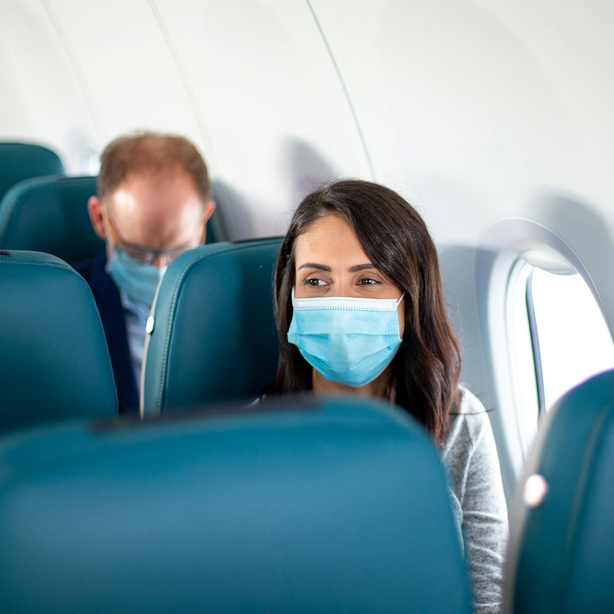 A lady with brown hair sitting on an airplane seat with a surgical mask on her face