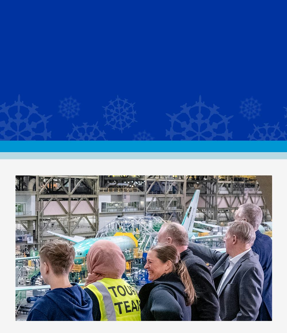 A group of people observes an airplane assembly inside a large hangar. A person in a yellow "Tour Team" vest is visible. The mood is curious and attentive.