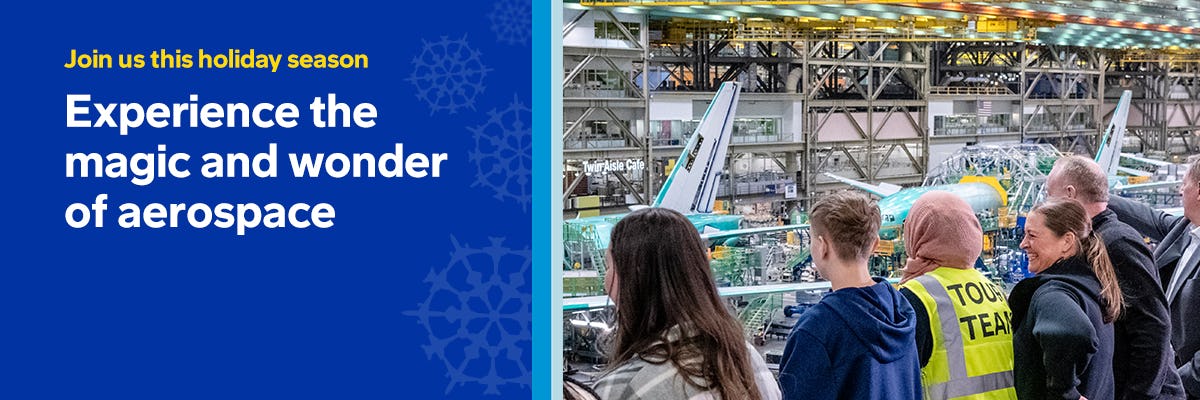 A group of people observes an airplane assembly inside a large hangar. A person in a yellow "Tour Team" vest is visible. The mood is curious and attentive.