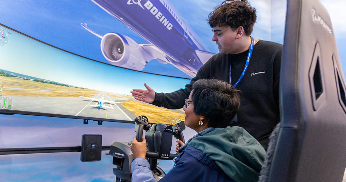 A person sits at a flight simulator, focused, while another stands nearby, gesturing towards the screen. The simulator displays a runway scene.