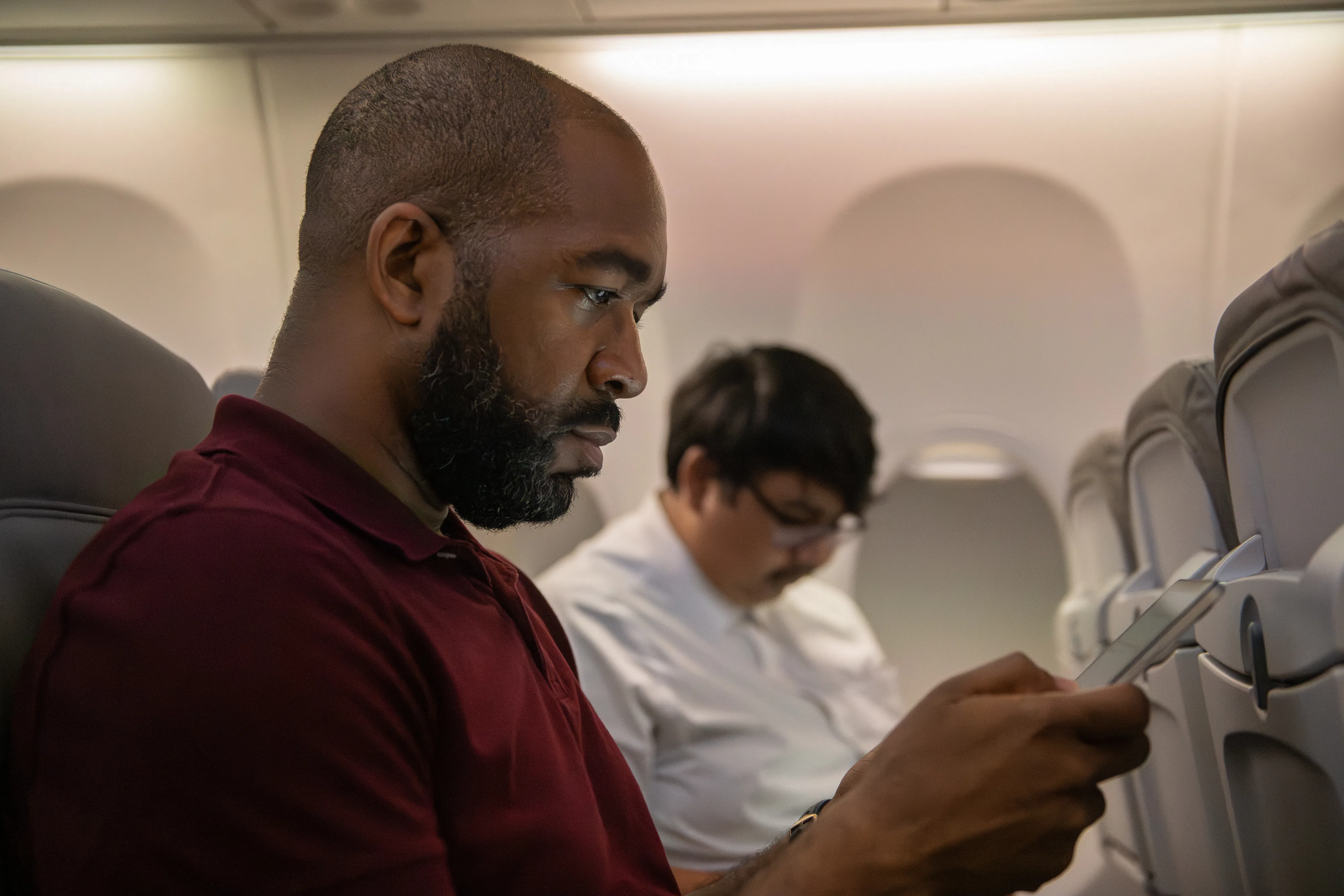 A man in a red shirt focuses intently on his phone while seated on an airplane. Another passenger in a white shirt sits behind, reading.