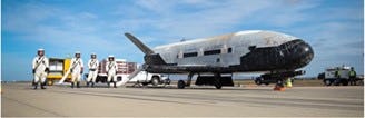 A shot of the X-37B next to three space travelers in full-body space suits from the tarmac.