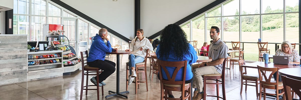 Guests sitting at tables in Paper Plane Café, some having conversations and others sitting alone with their laptop while bright light pours in through the floor-to-ceiling windows and our coffee/concessions bar off to the side.
